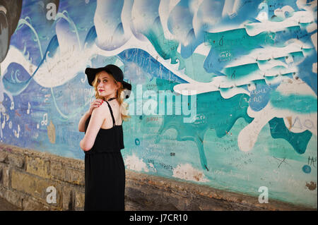 Blonde woman on black dress, necklaces and hat against graffiti wall. Stock Photo