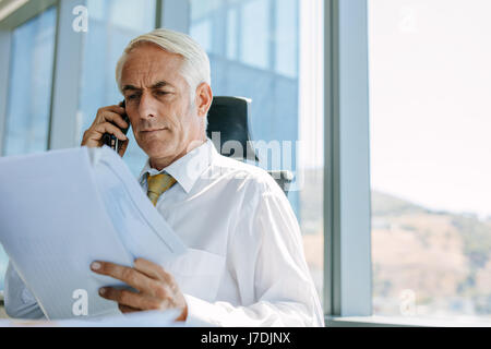 Shot of mature businessman sitting at his desk reading a document and talking on mobile phone. Senior male entrepreneur working at his office desk. Stock Photo