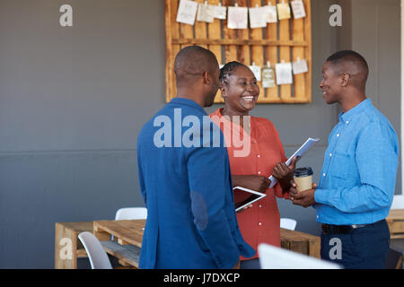Three casually dressed young African office coworkers smiling and talking together while standing in a large modern office Stock Photo