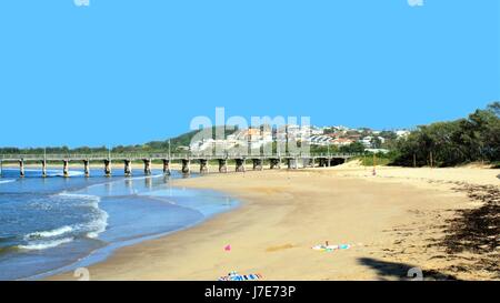 Jetty or Pier over sea with beach, sand and waves and unidentifiable people, hills and buildings in view. Coffs Harbour Jetty. Landscape of Australia. Stock Photo