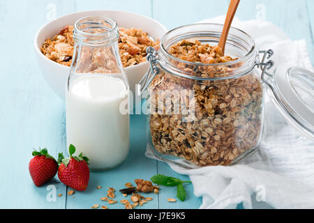 Homemade baked granola in a glass jar, a bottle of milk and strawberries on a turquoise wooden table. Healthy breakfast Stock Photo