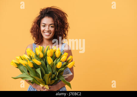 Happy African woman holding bouquet of flowers and looking at the camera over yellow background Stock Photo