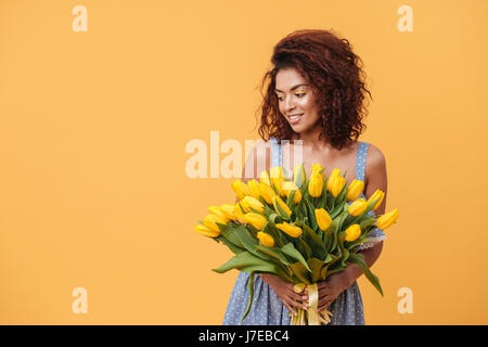 Smiling African woman holding bouquet of flowers and looking away over yellow Stock Photo