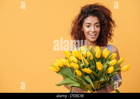 Surprised African woman holding bouquet of flowers and looking at the camera over yellow background Stock Photo