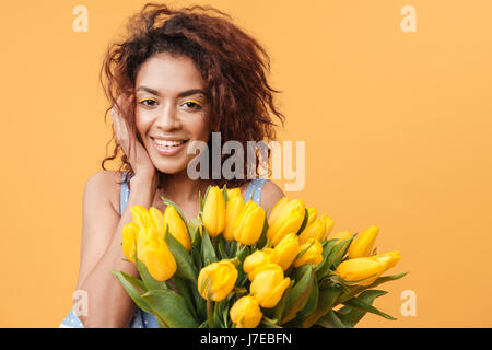 Cute African woman holding bouquet of flowers and looking at the camera over yellow background Stock Photo