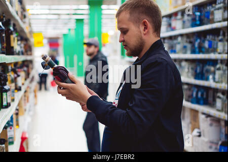 bearded man, In a black jacket, stopped to make my choice in store window, holds a bottle of strong alcohol in his hand, taking from shelf Stock Photo