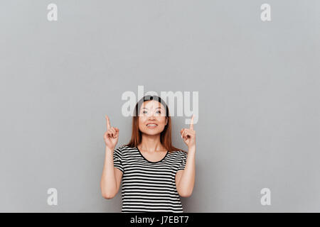 Picture of happy young asian woman standing over grey wall pointing. Looking aside. Stock Photo