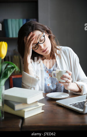 Portrait of a tired exhausted mature woman in eyeglasses leaning on her hand and holding cup of tea while working on laptop at home Stock Photo