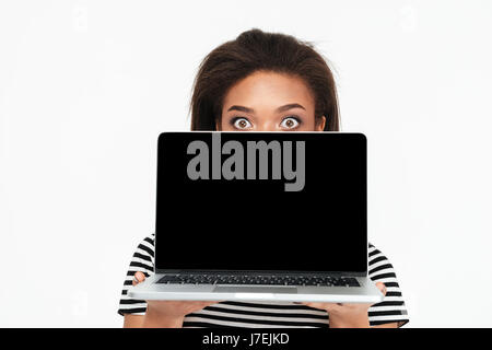 Portrait of a surprised shocked african woman hiding behind blank screen laptop isolated over white background Stock Photo