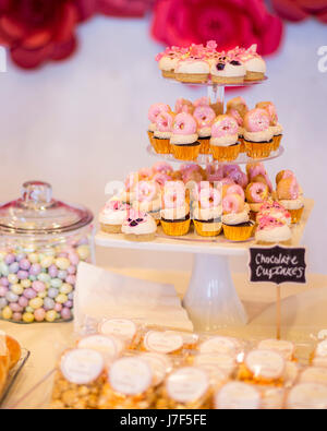 Mini chocolate cupcakes topped with mini pink donuts on a dessert table. They are displayed on a tiered display tray. There is a cute chalkboard sign. Stock Photo