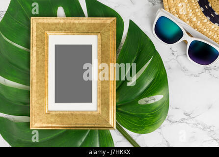 Blank Vintage golden color picture frame on green monstera leaf with sunglasses and straw hat on white marble table top view,Mock up for adding your p Stock Photo