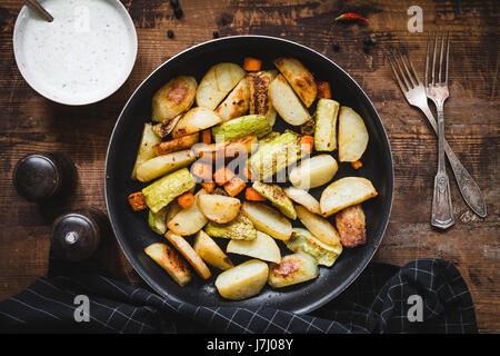 Roasted vegetables, potato wedges, carrots, onion, garlic and zucchini in pan with tzatziki sauce on rustic wooden table.