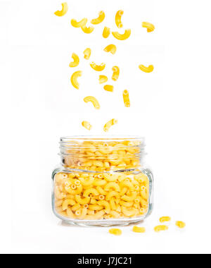 raw macaroni pasta in clear glass jar with pieces falling down into the jar and some on the ground, on isolated white background Stock Photo