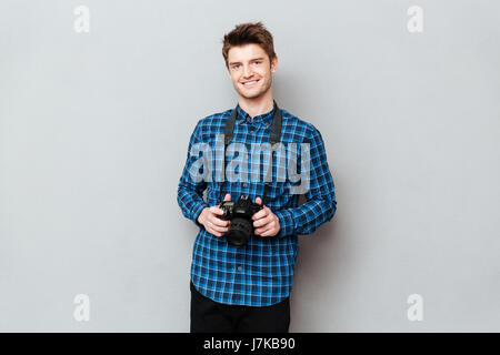 Young cute man holding camera in hands and smiling isolated over grey Stock Photo