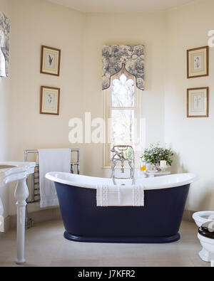 Dark blue freestanding bath in room with gothic windows and toile de jouy patterned pelmets Stock Photo