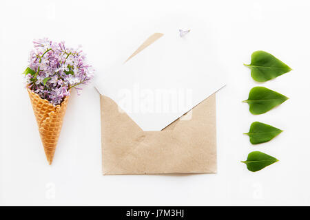 Composition of Bouquet of purple lilac flowers in the waffle cone, opened craft paper envelope with empty blank postcard and raw of green leafs on the Stock Photo