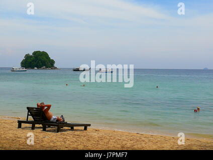 A single tourist relaxing at the beach in Tioman Island, Malaysia, on a deck chair while another have a swim in the beautiful blue sea. Stock Photo