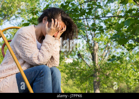 A young woman covering her face with both hands sitting on the top of a slide at a park, crying, suffering, Stock Photo