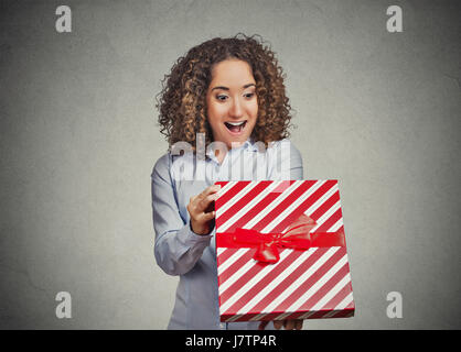 Closeup portrait happy super excited young woman opening unwrapping red birthday gift box, isolated grey wall background. Positive human emotions, fac Stock Photo