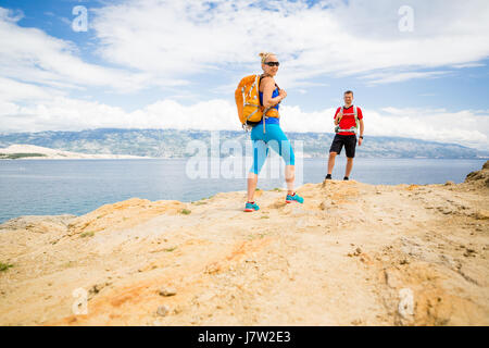 Couple hikers walking on trail at seaside. Man and woman hiking trekking in summer nature by a sea and mountains. Young couple on rocky mountain trail Stock Photo