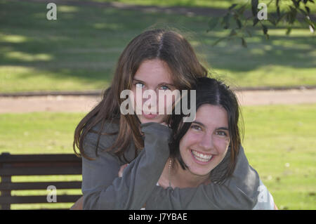 2 Teenage girls warm piggy back hug in shade on park bench with green background on sunny day Stock Photo