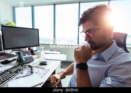 Close up of businessman wearing spectacles sitting in pensive mood  with hand to his chin. Young entrepreneur sitting at his work station looking down Stock Photo