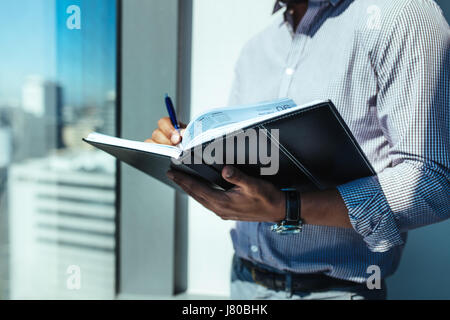 Businessman holding an open diary standing beside a window in office. Man doing office work standing in sunshine near window of highrise office buildi Stock Photo