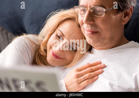 Happy middle aged couple sitting on bed, man reading newspaper at home Stock Photo
