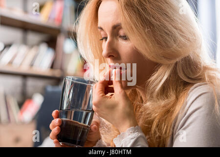 Blonde middle aged woman holding glass with water and taking pill Stock Photo