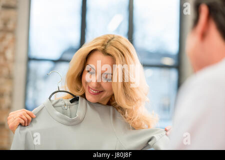 Happy blonde middle aged woman trying on grey fashionable dress indoors Stock Photo