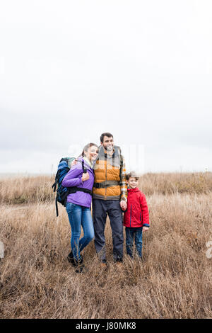 Happy family with backpacks standing in tall dry grass and looking away at cloudy autumn day Stock Photo