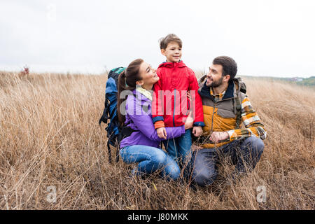 Happy family with backpacks embracing in tall dry grass and looking away at cloudy autumn day Stock Photo