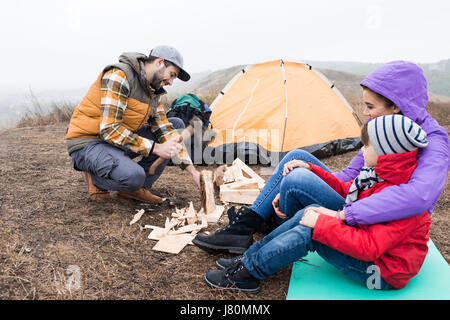 Side view of smiling mother embracing little son and looking at young man chopping firewood with axe Stock Photo