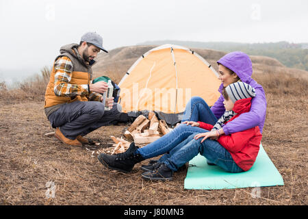 Side view of smiling mother embracing little son and looking at young man holding bottle with liquid for kindling fire. Happy young family sitting nea Stock Photo