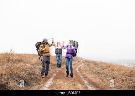 Happy young family with backpacks holding hands and walking on rural path at cloudy autumn day Stock Photo