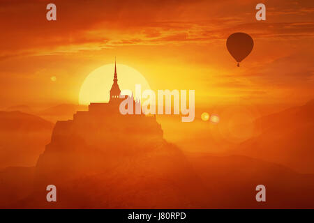 Beautiful sunset landscape over the misty kingdom between the orange hills in the center of nature and the silhouette of a flying air balloon. Fantasy Stock Photo