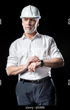 Mature male architect in white hard hat checking wristwatch on black Stock Photo