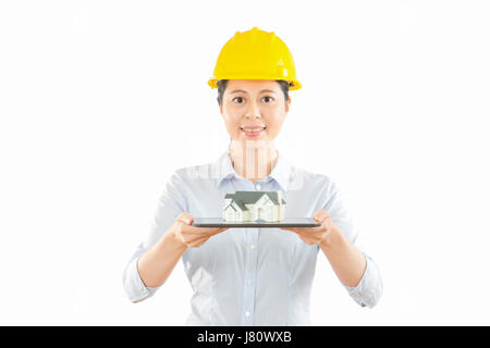 woman engineer of big blank background showing portable touch pad with miniature house model while standing in white wall with copyspace. concept of b Stock Photo