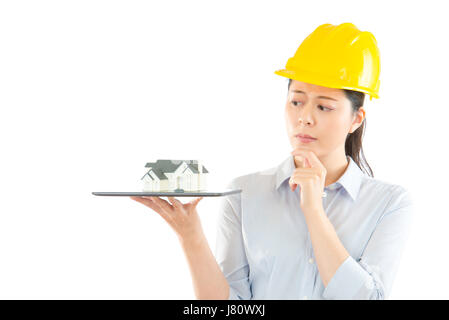 thoughtful confused builder in helmet thinking architecture design problem scratching his chin and holding pc tablet with house model isolated on a wh Stock Photo