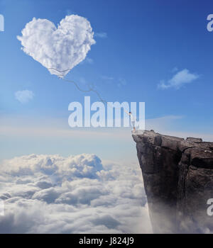 Imaginary view with a boy on the edge of a cliff, trying to catch a heart shaped cloud with a long rope. Follow your heart concept. Stock Photo