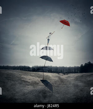 Young boy stand on a stack of umbrellas arranged as a ladder to the sky, trying to catch a different red umbrella. The pursuit of happiness and succes Stock Photo
