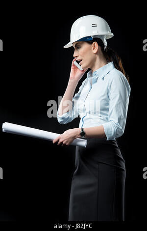 Young female architect in helmet holding blueprint and talking on smartphone Stock Photo