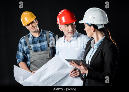 Two architects in hard hats chatting over blueprint while engineer waiting Stock Photo