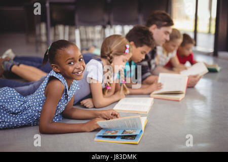 Teacher and schoolkids lying on floor while reading books in library at school Stock Photo