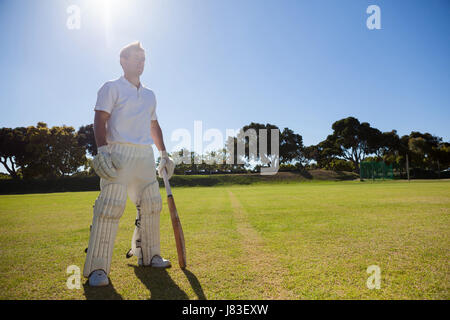 Full length of cricket player with bat standing on grassy field on sunny day Stock Photo