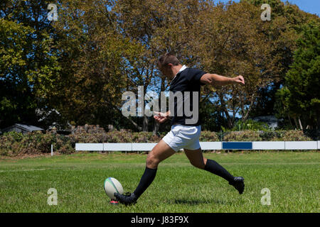 Determined rugby player kicking ball on grassy field against trees Stock Photo