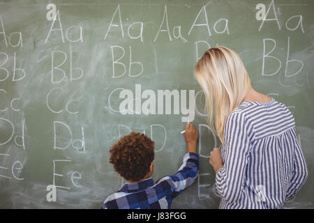 Teacher assisting schoolboy in writing alphabet on chalkboard at school Stock Photo