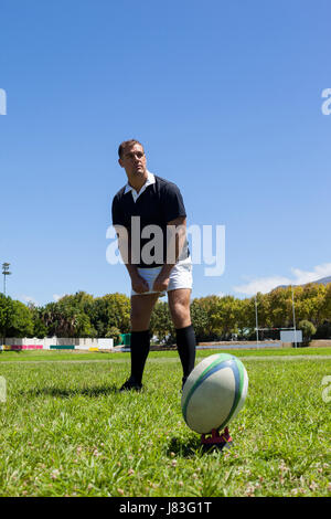 Rugby player looking away while standing by ball on grassy field against clear blue sky Stock Photo