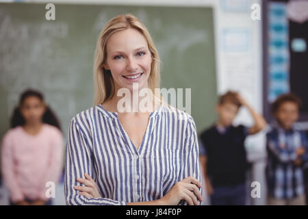 Portrait of happy teacher standing with arms crossed in classroom at school Stock Photo