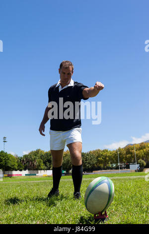 Full length of rugby player kicking ball on grassy field against clear blue sky Stock Photo